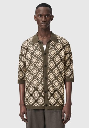 Homme portant un cardigan à mailles ouvertes marron et crème à motifs avec des boutons, col et manches vert foncé, sur un fond gris uni.