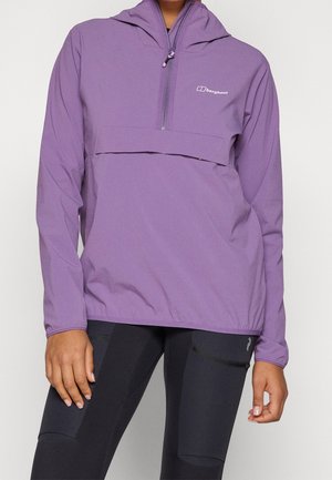 Person trägt eine lila Berghaus Halb-Reißverschluss-Kapuzenjacke und schwarze Leggings mit Reißverschlusstaschen, steht mit entspannten Händen an den Seiten.