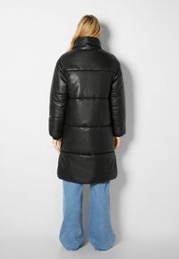 Černý vatovaný kabát vyrobený z kůže, s vysokým límcem, dlouhými rukávy a střední délkou. Kombinovaný s modr ými džíny širokého střihu.