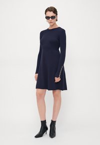 Abito lungo a maniche lunghe in navy con orlo svasato, dotato di dettagli con bottoni sulle maniche, abbinato a stivali neri alla caviglia.
