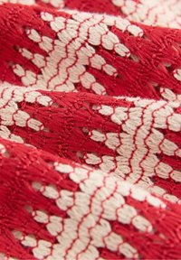 Tela bordada en rojo y crema con un patrón texturizado que presenta formas alternas. Textura suave con costuras visibles y detalles intrincados.