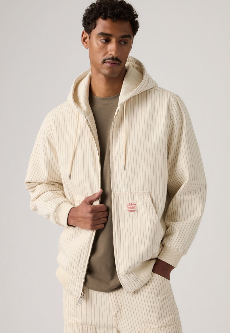 Άνδρας που φοράει κρεμ χρωματιστό φούτερ με ρίγες και ταιριαστό παντελόνι, με πουκάμισο χακί από μέσα, στέκεται μπροστά σε απλό ανοιχτό φόντο.
