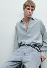 Chemise à manches longues gris clair avec une coupe décontractée et un col, associée à un jean en denim bleu clair maintenu par une ceinture tissée noire. Détails en tissu texturé.