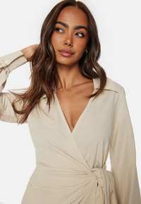 Beige omslagklänning med djup V-ringning, långa ärmar och knytning i midjan. Mjuk, lättviktsmaterial med en slät struktur.