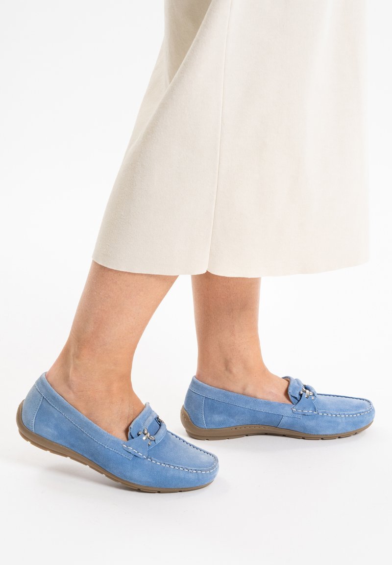 Blauwe suède instappers met een laag profiel, decoratieve band en gestikt detail. Rubberen zool biedt grip. G gedragen met een lichtbeige rok.