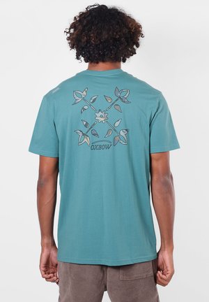 Homme aux cheveux bouclés portant un t-shirt bleu sarcelle avec un motif de fleurs et feuilles croisées avec le texte "OXBOW" au dos, debout devant un fond uni.