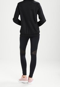 Svart zip-up jacka, åtsittande svarta leggings med nätaccent och ljusrosa träningsskor. Slät tyg, smalnande design och elastiskt material.