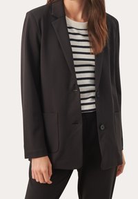 Černý látkový blazer s klopovým límcem, předními knoflíky a postranními kapsami, nošený přes pruhované tričko. Hladká textura a přiléhavý střih.