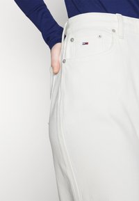 Jeans de mezclilla blanca con herrajes metálicos y un pequeño logotipo bordado. Presenta un corte recto y bolsillos laterales. Textura suave.