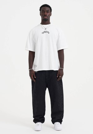 White oversized t-shirt with black logo on chest, paired with loose black jeans featuring a subtle pattern. White sneakers complete the outfit.