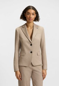 Blazer beige su misura con una texture liscia, rever a punta, due bottoni neri e un logo discreto sulla tasca del petto.