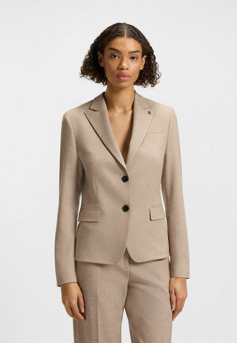 Blazer beige su misura con una texture liscia, rever a punta, due bottoni neri e un logo discreto sulla tasca del petto.