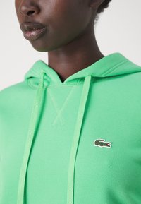 Felpa verde con tessuto testurizzato, coulisse regolabile e logo ricamato sul petto. Inquadratura ravvicinata che mette in evidenza il design del collo e del cappuccio.