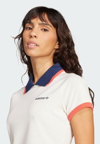 Biele polo tričko s tmavomodrým a korálovým golierom, krátkymi rukávmi a vyšívaným logom Adidas na ľavej hrudi. Klasický dizajn.