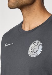 Maglietta in cotone grigia con logo rotondo del Paris Saint-Germain e il logo Nike argento sul lato sinistro del petto. Design con girocollo semplice.