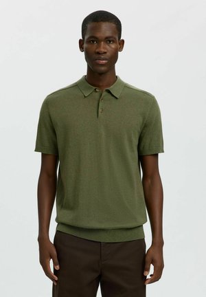 Jonge man die een olijfgroen poloshirt met korte mouwen en donkerbruine broek draagt, staand tegen een effen lichte achtergrond.