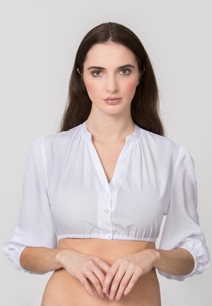 Young woman with long dark hair wears white cropped blouse with buttons, standing against a plain light gray background.
