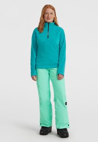 Maglione in pile turchese con zip a metà e dettagli neri, abbinato a pantaloni da neve verde chiaro. Il modello indossa stivaletti da neve neri.