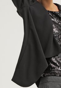 Czarna asymetryczna marynarka noszona na czarnej topie z cekinami, w połączeniu z szarymi spodniami, pokazana od talii do szyi.