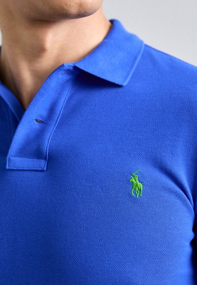 Modra polo majica iz teksturirane tkanine z zelenim vezenim logotipom konja in jahača na levi prsni strani. Ima klasičen ovratnik.