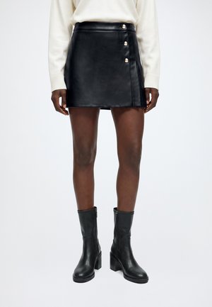 Mini-jupe noire en simili cuir avec trois boutons décoratifs, associée à des bottines noires à talons et un top blanc à manches longues.