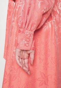 Korallfärgad klänning med blommönster i jacquard, med puffärmar och en knappmanschett med tre matchande knappar. Slät textur.