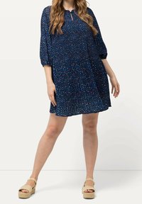 Kvinna iklädd en marinblå knälång klänning med små flerfärgade blommönster och beige kilklackar, stående med benen isär.