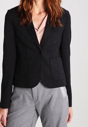 Kobieta ubrana w dopasowaną czarną marynarkę na różowej bluzce, w parze z szarymi dopasowanymi spodniami, stojąca na jasnym, jednolitym tle.