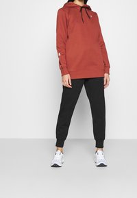 Rostfärgad huvtröja med justerbar snörning, bärs över svarta tapered joggers och vita sportskor. Minimalistisk design.