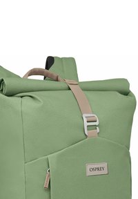 Sac à dos enroulable vert en matériau texturé, avec une bandoulière ajustable beige et une poche avant zippée. Patch logo visible.
