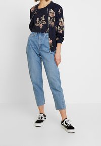 Námořnická květinová bomber bunda přes černý top, v kombinaci s vysokými světle modrými džíny a černými teniskami s bílými detaily.