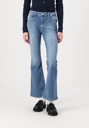 Person iført lyseblå flare jeans, sort langærmet top, hvide sokker og sorte loafers med kvaster mod en ensfarvet hvid baggrund.