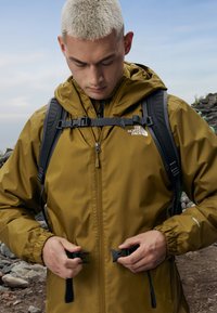 Hnedá nepremokavá bunda s kapucňou, nastaviteľným pásom a predným zipsom. Logo The North Face je viditeľné na hrudi. Ruksak nosený na pleciach.