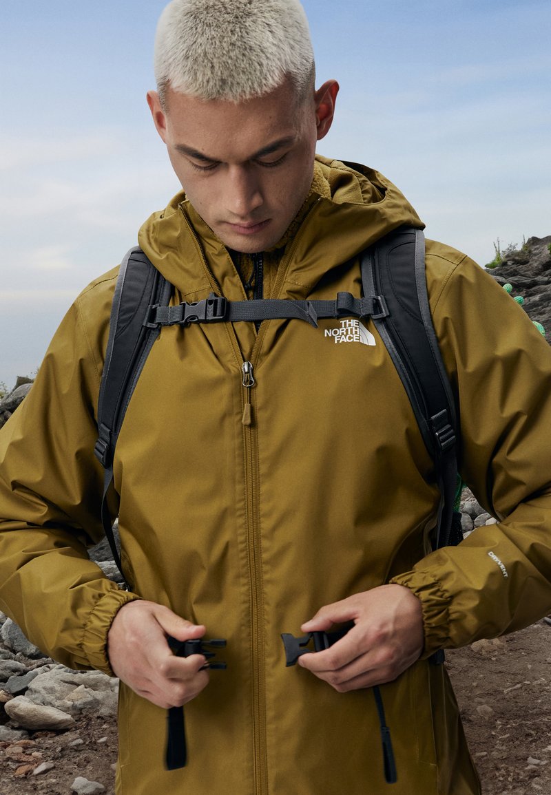 Hnedá nepremokavá bunda s kapucňou, nastaviteľným pásom a predným zipsom. Logo The North Face je viditeľné na hrudi. Ruksak nosený na pleciach.