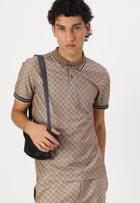 Braun gemustertes Poloshirt mit einem Kragen und kontrastierendem schwarz-weißem Saum. Das Model hält eine kleine schwarze Tasche.