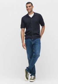 Korte mouwen marineblauw overhemd met een gemêleerd patroon, gecombineerd met blauwe jeans en groene sneakers. Model staat in een ontspannen houding.