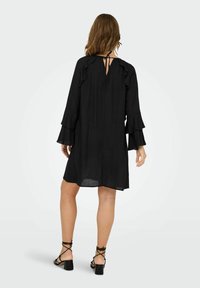 Vestido negro con mangas acampanadas, escote redondo en la parte posterior, cierre con lazo y diseño hasta la rodilla. Llevado con tacones negros con tiras.