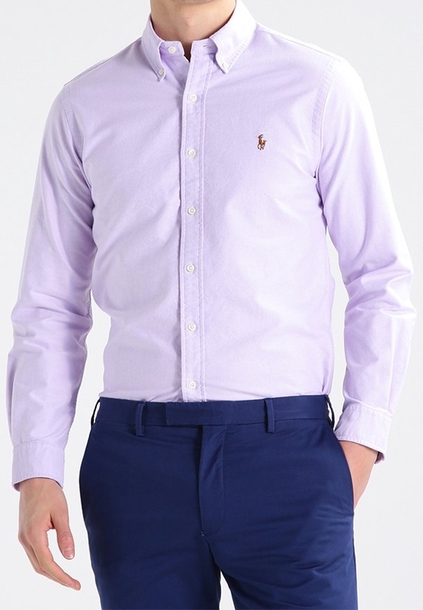 Homme portant une chemise violette claire à boutons avec un petit logo brodé sur la poitrine et un pantalon bleu foncé, une main dans la poche.