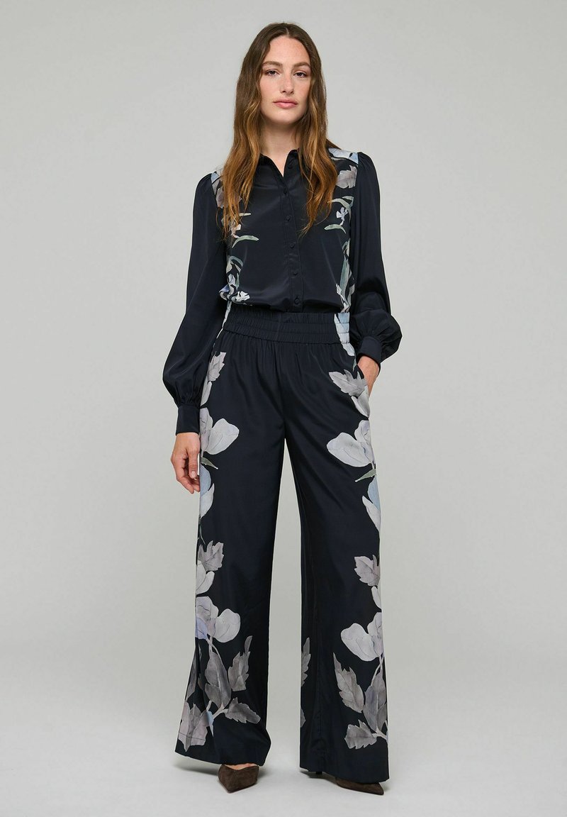 Femme portant un chemisier noir boutonné et un pantalon large assorti avec de grandes imprimés floraux gris, debout avec une main dans la poche.