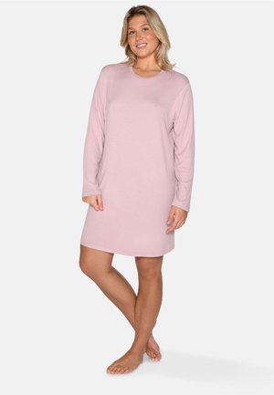 Femme debout pieds nus, vêtue d'une robe côtelée rose pâle à manches longues, souriant avec une posture détendue sur un fond uni.