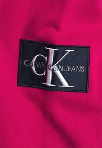 Calvin Klein Jeans logo patch op een felroze stof. De patch is zwart, met witte borduursels met de merknaam en initialen.