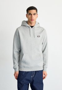 Felpa grigia in cotone misto con tasca frontale, cappuccio regolabile con cordino e patch del marchio sul lato sinistro del petto. Indossata con jeans scuri.