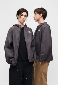 Δύο νεαροί ενήλικες φορούν γκρι καρό μπουφάν North Face, ο ένας κοιτάζει μπροστά με τα χέρια στις τσέπες, ο άλλος προφίλ.