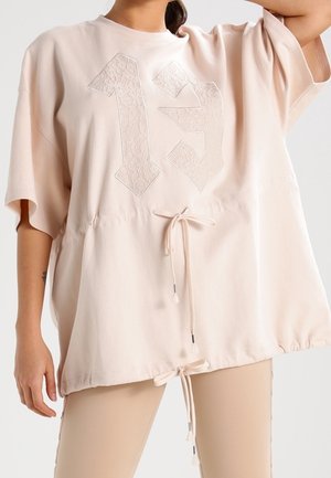 Top suelto de manga corta en rosa pálido con el número 13 en patrón de encaje en el frente, con cordones en la cintura y el dobladillo, combinado con leggings beige.