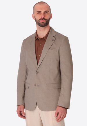 Uomo con la barba che indossa un blazer beige sopra una camicia marrone e pantaloni chiari, in piedi davanti a uno sfondo semplice.