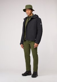 Giacca impermeabile nera con cappuccio, camicia verde con bottoni, pantaloni cargo verdi e scarpe da trekking nere con dettagli arancioni.