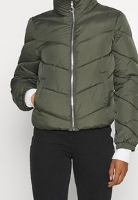 Giacca bomber imbottita verde con collo alto, chiusura a zip, polsini bianchi a coste e motivo a quilting a chevron.