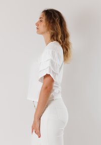 T-shirt bianco corto con maniche corte a volant, abbinato a jeans bianchi. Texture liscia, design semplice e senza motivi o accessori visibili.