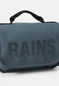 Modrý vodoodolný vak s matným povrchom, vybavený čiernym popruhom a reliéfne vyrazeným logom "RAINS" na prednej strane. Hladká textura a obdlžnikový tvar.