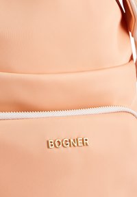 Mochila de tela melocotón con una textura suave, que presenta un logo dorado "BOGNER" y un acento de cremallera blanca. Diseño sencillo y forma práctica.